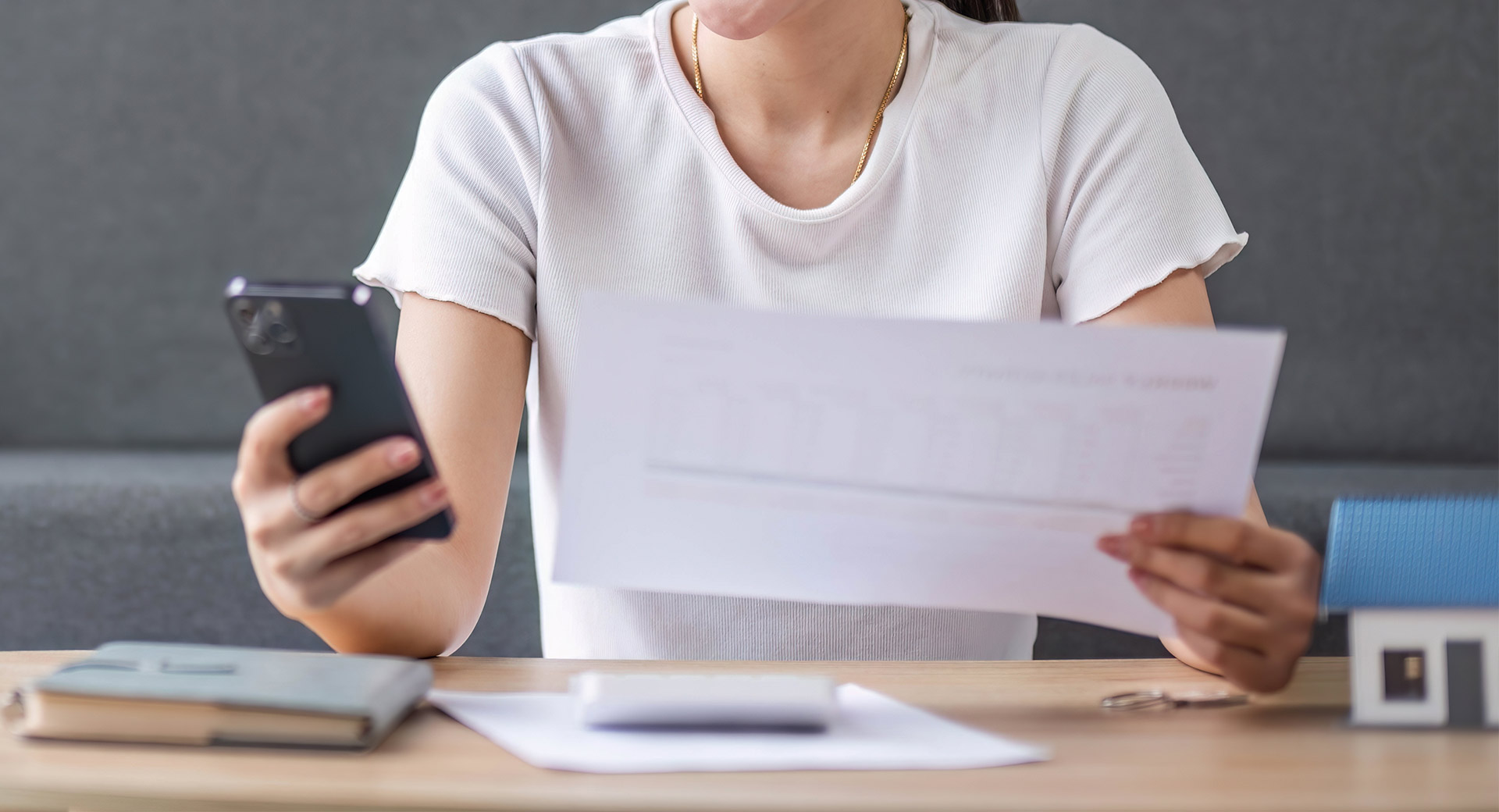 A person sitting at a desk holds a bill in one hand and a mobile phone in the other